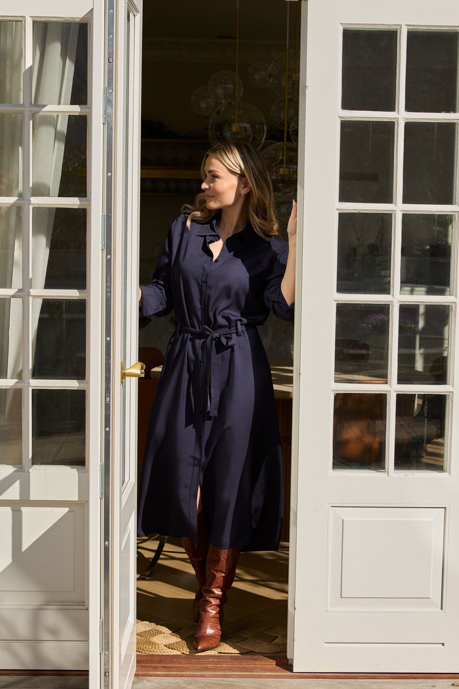 Kvinde iført tidløs skjortekjole i navy fra A Working Theory, designet i blød viskose med bindebånd i taljen. Klassisk og elegant skjortekjole til kvinder, vist i døråbning med naturligt lys – velegnet til både arbejde og hverdagsbrug.